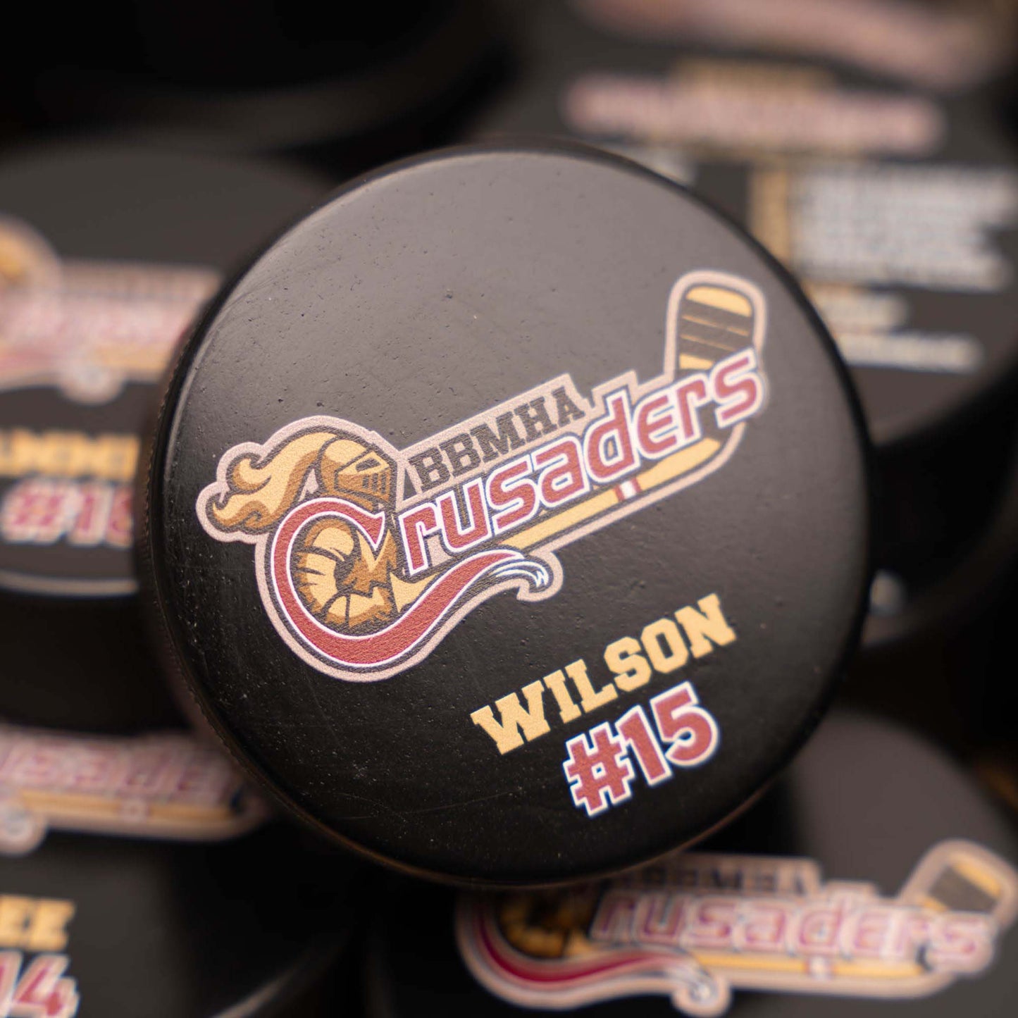 Hockey puck with 'BBMHA Crusaders' and 'Wilson #15' branding on a blurred background.